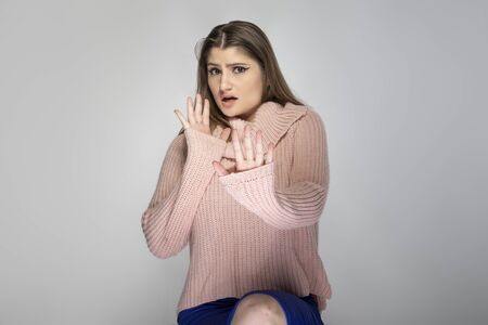 Close up portrait of a young Caucasian woman wearing a pink sweater on a grey background.  The model is holding hands up as a stop gestureの写真素材