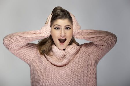 Close up portrait of a young Caucasian woman wearing a pink sweater on a grey background.  The model looks surprised and amazedの写真素材