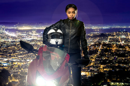 Sporty female motorcyclist or biker wearing a leather jacket and helmet on a road at night with a motorcycle above a scenic view of the San Francisco cityscapeの写真素材