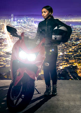 Sporty female motorcyclist or biker wearing a leather jacket and helmet on a road at night with a motorcycle above a scenic view of the San Francisco cityscapeの写真素材