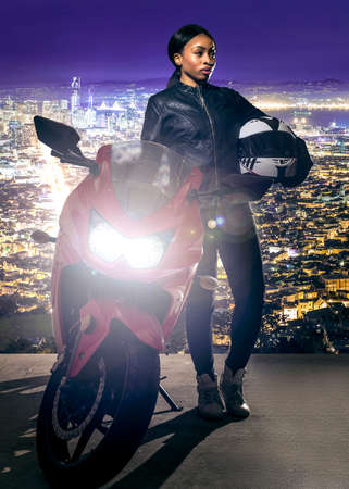 Sporty female motorcyclist or biker wearing a leather jacket and helmet on a road at night with a motorcycle above a scenic view of the San Francisco cityscapeの写真素材