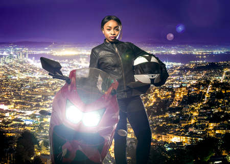 Sporty female motorcyclist or biker wearing a leather jacket and helmet on a road at night with a motorcycle above a scenic view of the San Francisco cityscapeの写真素材