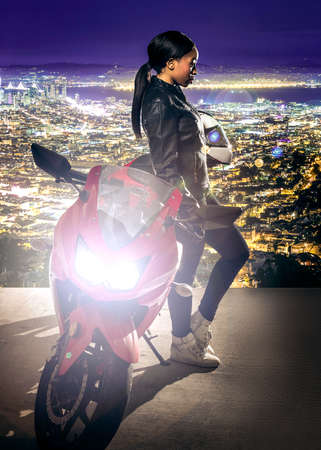 Sporty female motorcyclist or biker wearing a leather jacket and helmet on a road at night with a motorcycle above a scenic view of the San Francisco cityscapeの写真素材