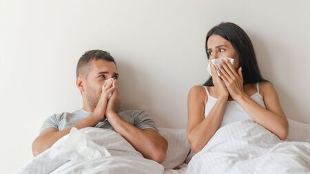 Sick couple feeling unwell together because of getting cold. Man and woman look on each other while sneezingの写真素材