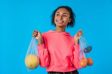 Happy african american woman smiling and carrying net bag with fruits isolated over blue backgroundの写真素材