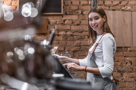 Portrait of happy young woman at counter with cashbox working in coffee shop