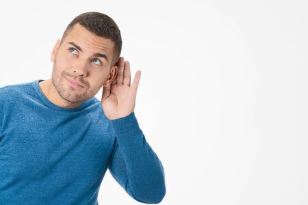 Curious young man keeping hand at his ear while overhearing secret over white background