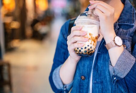 Young girl in denim jacket is drinking brown sugar flavored tapioca pearl bubble milk tea with glass straw in night market of Taiwan, close up, bokehの写真素材