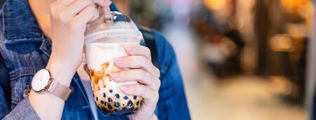 Young girl in denim jacket is drinking brown sugar flavored tapioca pearl bubble milk tea with glass straw in night market of Taiwan, close up, bokehの写真素材