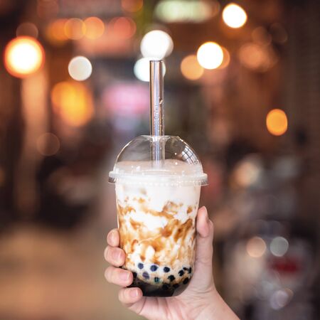 Young woman is holding, drinking brown sugar flavored tapioca pearl bubble milk tea with glass straw in night market of Taiwan, close up, bokehの写真素材