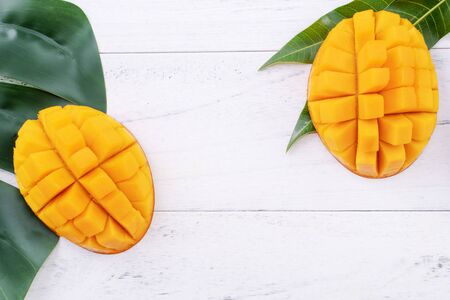 Beautiful chopped ripe mango with green leaves on bright white color wooden background, top view, flat lay, copy space. Tropical fruit design concept.の写真素材