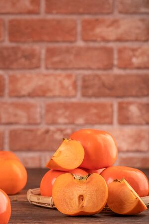 Sliced sweet persimmon kaki in a bamboo sieve basket on dark wooden table with red brick wall background, Chinese lunar new year fruit design concept, close up.の写真素材