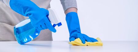 Young woman housekeeper in apron is cleaning, wiping down table surface with blue gloves, wet yellow...