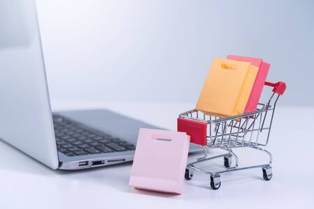 Online shopping. Mini shop cart trolley with colorful paper bags over a laptop computer on white table background, buying at home concept, close upの写真素材