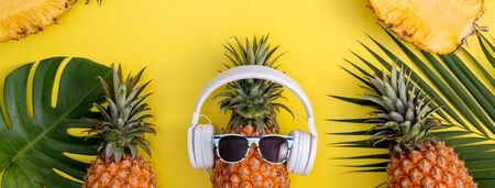 Funny pineapple wearing white headphone, listen music, isolated on yellow background with tropical palm leaves, top view, flat lay design concept.の写真素材