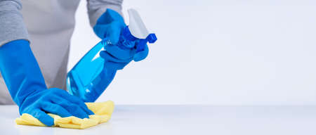 Young woman housekeeper in apron is cleaning, wiping down table surface with blue gloves, wet yellow rag, spraying bottle cleaner, closeup design concept.の写真素材