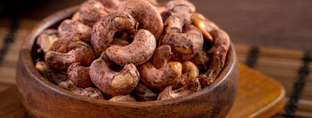 Cashew nuts with peel in a wooden bowl on wooden tray and table background, healthy raw food plate.