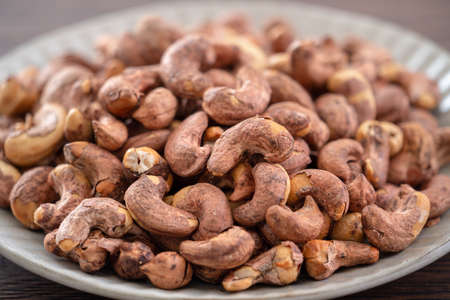 Cashew nuts with peel in a plate on wooden tray and table background, healthy raw food concept.の写真素材