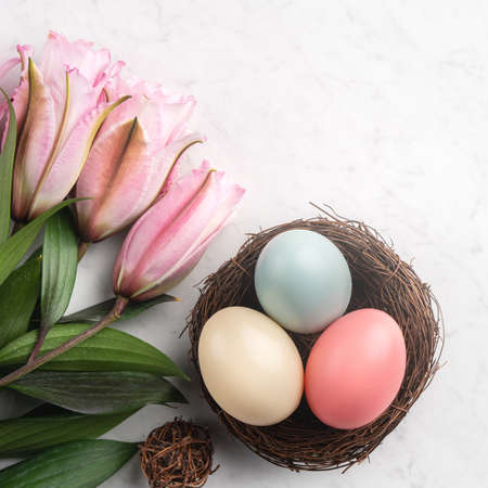 Colorful Easter eggs in the nest with pink lily flower on bright marble white table background.