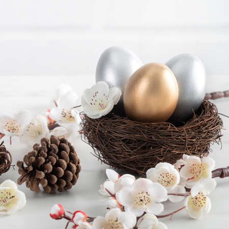 Close up of golden and silver Easter eggs in the nest with white plum flower on bright white wooden table background.の写真素材
