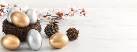 Close up of golden and silver Easter eggs in the nest with white plum flower on bright white wooden...
