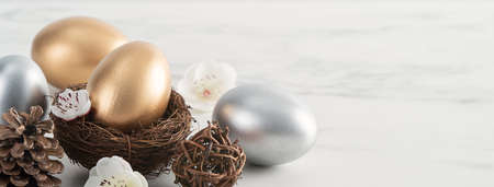 Close up of golden and silver Easter eggs in the nest with white plum flower on bright white wooden table background.の写真素材