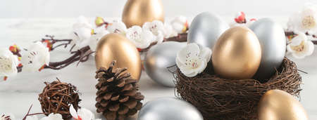 Close up of golden and silver Easter eggs in the nest with white plum flower on bright white wooden table background.の写真素材