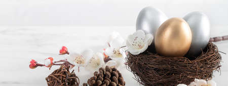 Close up of golden and silver Easter eggs in the nest with white plum flower on bright white wooden...