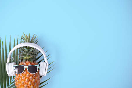 Funny pineapple wearing white headphone, concept of listening music, isolated on blue background with tropical palm leaves, top view, flat lay design.の写真素材