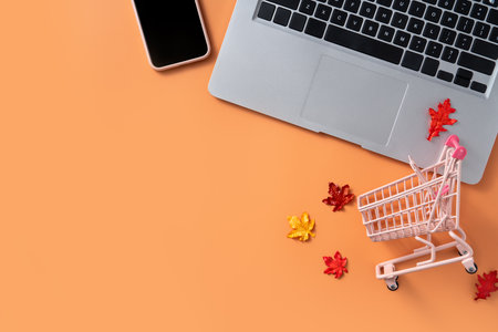 Autumn online shopping design concept with shopping cart, laptop, smart phone and maple leaves on orange table background.の写真素材