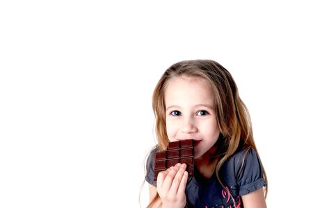 Cute blond smiling little girl eating chocolate tablet with tricky expression, isolated on white, space for textの写真素材