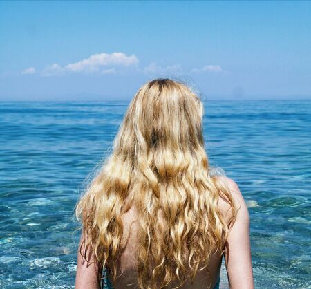 Beautiful girl with long blond hairs is sitting on the beach and looking at the sea.