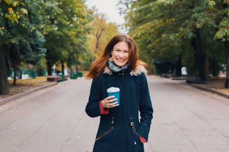 Redhead happy woman walking through the park alley, holding a blue paper cup of coffee or tea. Resting after work and looking at camera woman enjoying outdoors in autumn nature backgroundの写真素材