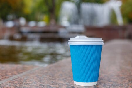 One blue paper coffee cup standing on granite surface in the city park on fountain background. Clouse up.の写真素材