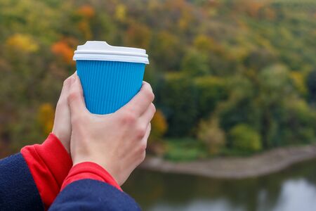 Close-up a woman hands holding a blue paper cup of coffee on autumn nature background with colorful trees and river.の写真素材