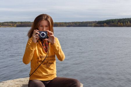 Young girl in yellow hoodie taking photo sitting on stone near river shore. healthy lifestyle, travel, weekend trip conceptの写真素材
