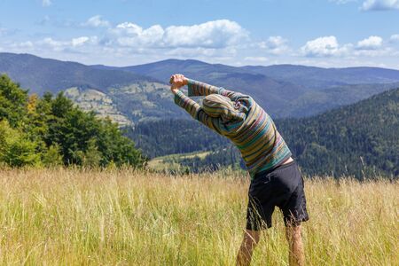 Back view of young man in green hoodie, exercising and stretching hands, looking at mountains range, resting after climbing and hiking on summer day - healthy lifestyle, travel, weekend trip conceptの写真素材