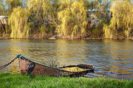 Old rusty metal boat chained to the river bank on a sunny autumn day. There is a water in the boat and the sun is gold reflected in itの写真素材