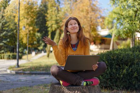Young girl in yellow hoodie with laptop, looking up, expressing surprise emotions and wondering outdoors, sitting on stump in autumn parkの写真素材