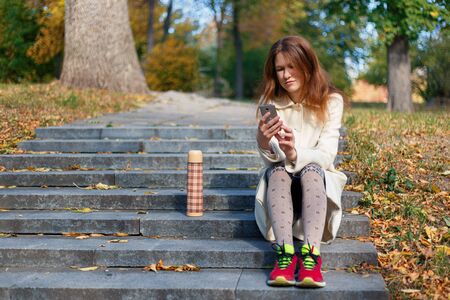 Sad young caucasian woman looking at mobile phone on the stairs in the autumn park outdoors. Thinking boring redhead girl upset reading bad newsの写真素材
