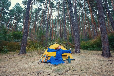 Young pretty redhead girl tourist in blue robe stretching hands after wake up, doing exercises, sitting in yellow tent, relaxing, enjoying nature in autumn forest. Travel weekend outdoors conceptの写真素材