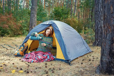 Young redhead girl pouring tea in cup from thermal flask, sitting in grey tent in autumn forest. Hiking, travel, weekend trip concept. Healthy active lifestyleの写真素材