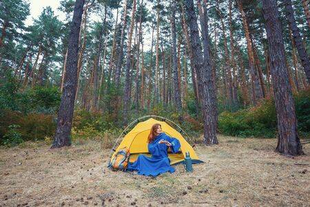 Young pretty redhead girl tourist in blue robe stretching hands after wake up, doing exercises, sitting in yellow tent, relaxing, enjoying nature in autumn forest. Travel weekend outdoors conceptの写真素材
