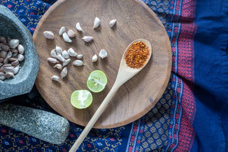 Thai cooking kitchen concept, garlics in stone mortar beside with pestle and piece of lime and dried chilly on traditional cloth backgroundの写真素材