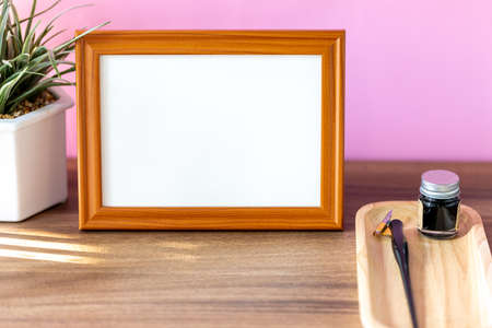 Blank landscape wooden frame beside a small houseplant and calligraphy tools, painting or artwork display mockup in front of pink wallの写真素材