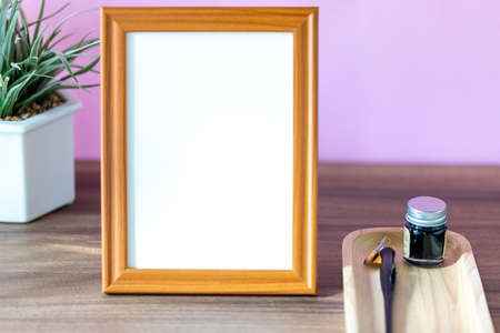 Blank vertical wooden frame beside a small houseplant and calligraphy tools, painting or artwork...