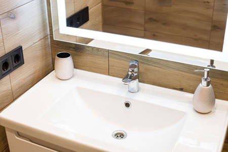 Modern washbasin with chrome faucet beside a stylish soap dispenser. Mirror with built-in led lighting.の写真素材