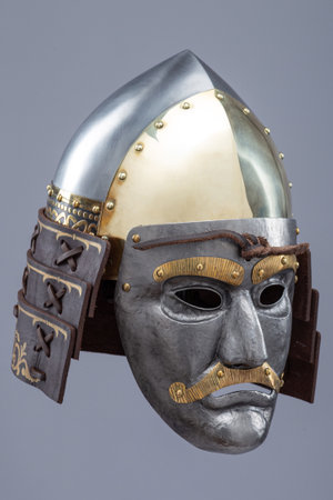 Historical metallic combat helmet with integrated face mask. Pointed steel helmet featuring a detailed metallic face mask with eye openings, decorative elements, and side protection flaps.の写真素材