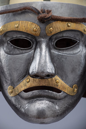 Historical metallic combat helmet with integrated face mask. Pointed steel helmet featuring a detailed metallic face mask with eye openings, decorative elements, and side protection flaps.の写真素材