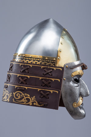Historical metallic combat helmet with integrated face mask. Pointed steel helmet featuring a detailed metallic face mask with eye openings, decorative elements, and side protection flaps.の写真素材